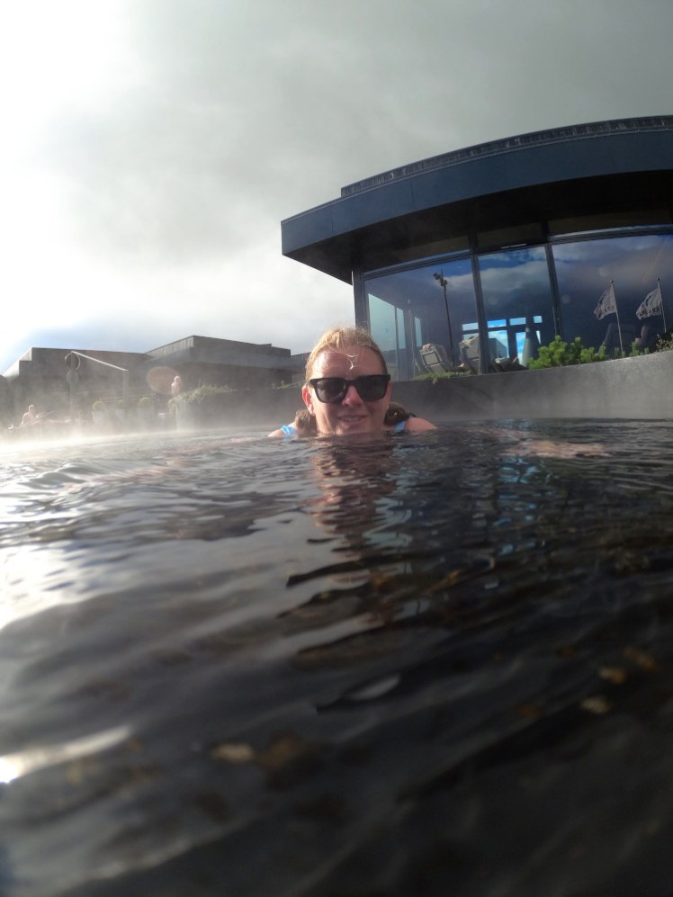 A GoPro selfie in a shallow black hot pool with more steaming pools in the background and an angular black building with floor-to-ceiling windows.