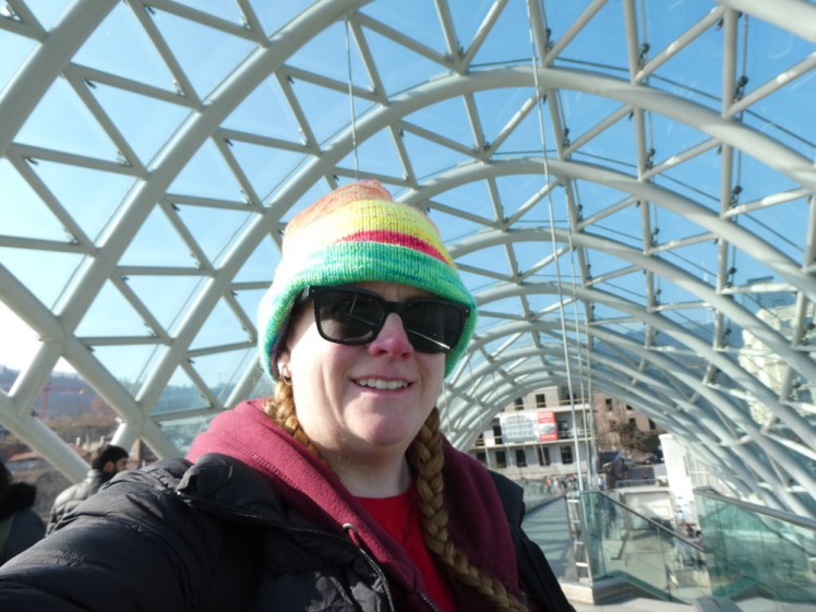 A selfie on the Peace Bridge. I'm wearing a neo stripy hat and the bridge canopy of criss-crossing steel tubes is above me.