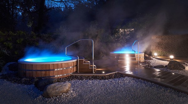 Two outdoor hot tubs with wooden decorative outsides like giant barrels, glow blue and steam in the dark evening chill.