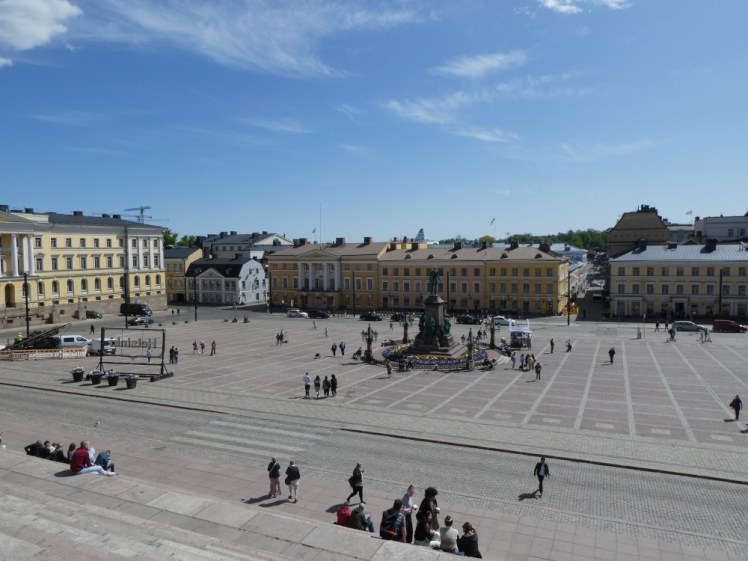 Senate Square in the sunshine, a cobbled square surrounded by yellow neoclassical buildings, as seen from the steps leading up to the cathedral.