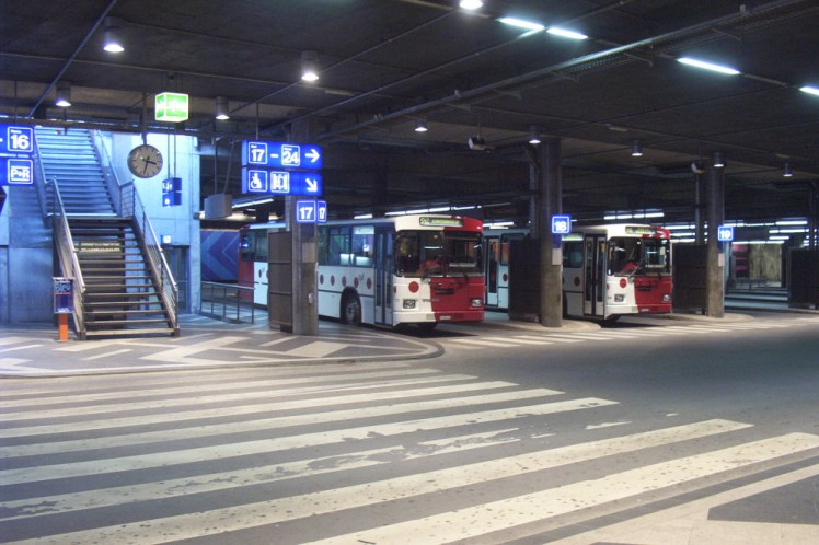 Fribourg bus station, which is underground, judging by the stairs coming down the left with daylight visible at the top. There are three stands visible and at two of them is a single-decker bus, half white, half dark red with red spots on the white parts.