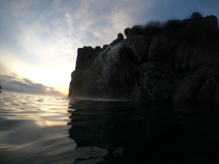 The Sky Lagoon seascape, with a cliff to the right and a waterfall tumbling over it. To the right, the sun is low and glowing brightly, reflected in the lagoon's water.