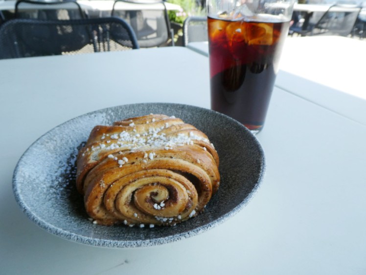 A large cinnamon roll in a shallow bowl. Its spirals are really well-defined and there's also a pint glass of Coca-Cola next to it.