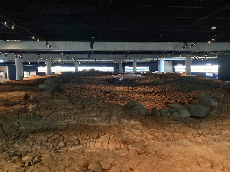 Looking up the longhouse, which can be faintly seen marked by lines of bigger darker stones among the reddish gravel. In the middle, a fire is projected onto an angled piece of plastic to show what it would have looked like inside.