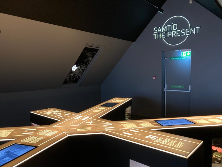 A model of Reykjavik Domestic Airport's runways, with info and screens set into it. The model is in a dark blue room with a sloping ceiling coming a very long way down and the words SAMTID, THE PRESENT in neon letters above a fire escape.