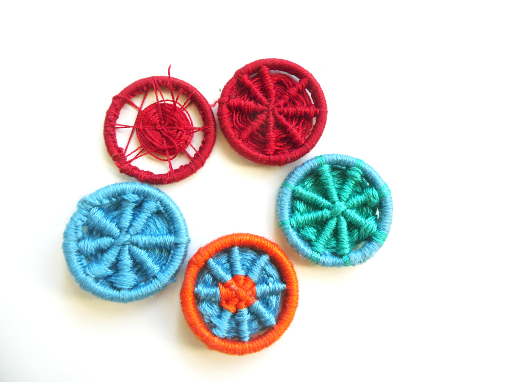 Five Dorset buttons in a circle. The first is in dark red and very gappy. The others are full. One is dark red, one is light blue with green spokes, one is orange with blue spokes and an orange centre and the last is all light blue.