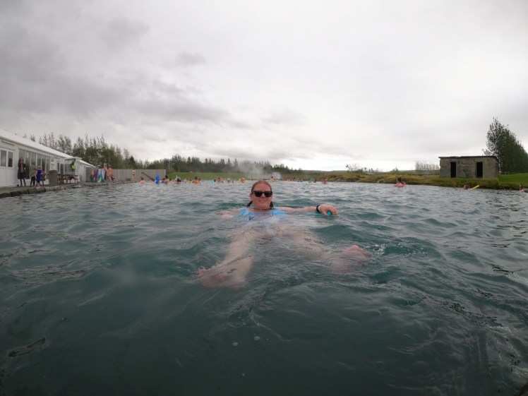 A timer selfie in the Secret Lagoon. I'm floating on my back with feet pointed towards the camera and a blue pool noodle under my arms. The water is blue and relatively translucent and the sky is the heavy grey of it being about to rain.