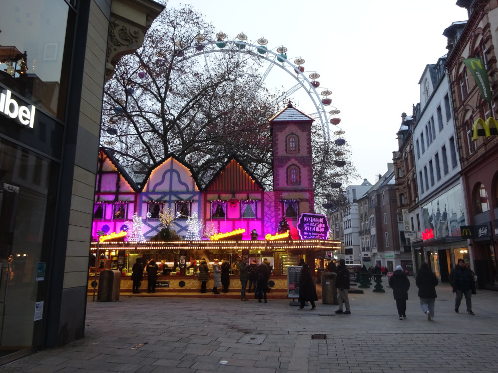 A yellow-lit bar in the middle of the street. The top of the bar is set up to look like a Hanseatic street scene with pink lights. Behind it is a ferris wheel.