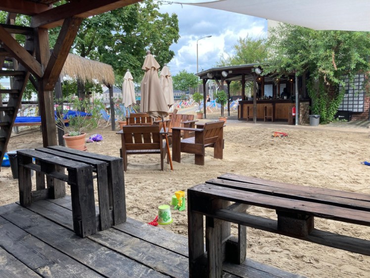 An artificial beach by the river in Berlin - a sand-covered area with wooden furniture and large shady umbrellas, currently furled. On the other side of a path is a bar with a lot of greenery hanging over it.