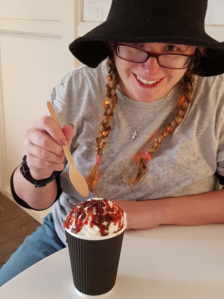 Me with a small wooden spoon about to poke my Haunted Forest Hot Chocolate. It's in a takeaway cup, topped with cream, chocolate flakes and cherry sauce. I've taken off my blanket cardigan but not the hat and the close-up means you can now see I have tiny orange fairy lights threaded through my plaits.