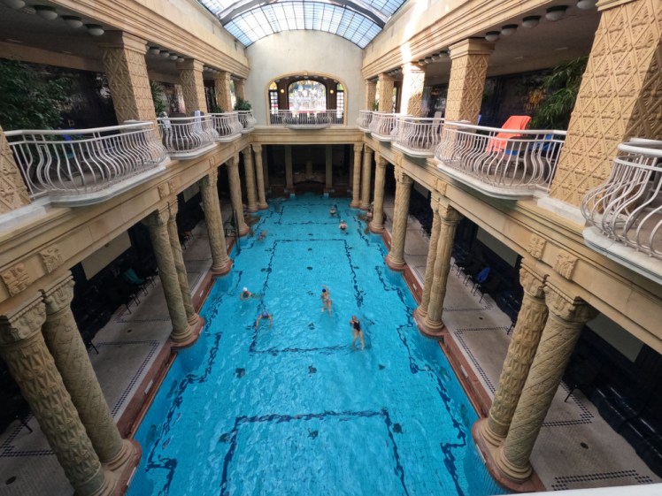 The pool at Gellert, a beautiful blue pool surrounded by yellow stone columns leading up a balcony around the pool and white iron balconettes for overlooking the pool.