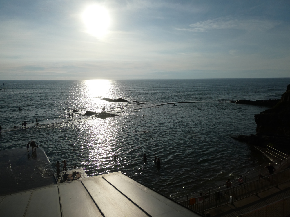 Navy blue open sea with a low sun reflecting on it - but if you look closely, among the rocks sticking out of the water, you can see a concrete wall enclosing a pool area.