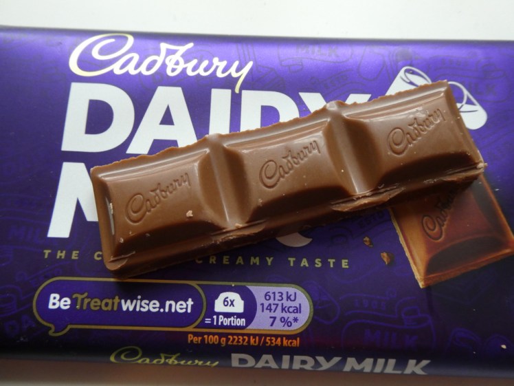 A bar of Dairy Milk chocolate with three squares lying on top of the purple packaging.