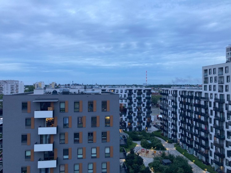 The view from my Warsaw hotel window as the sky turns the mid-blue of evening. In the foreground are a lot of apartment buildings but there are trees and countryside and a power station if you look beyond them.