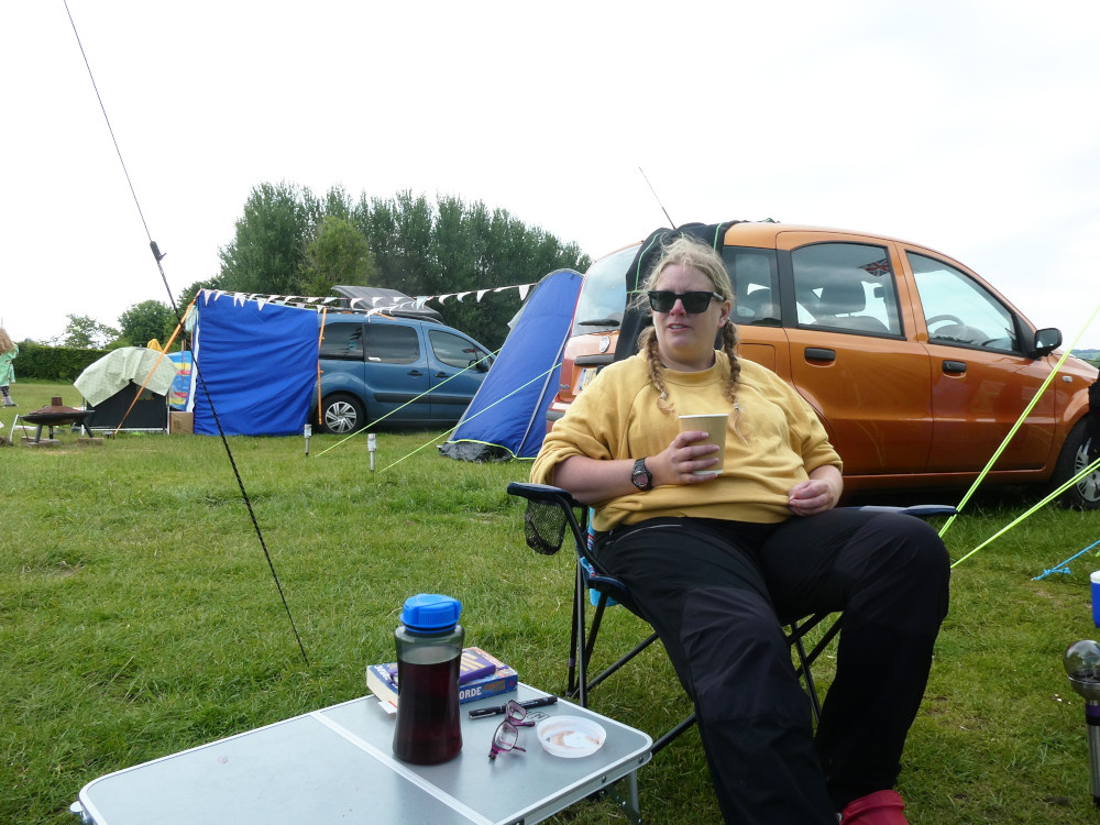 A timer selfie, sitting in a camp chair outside my tent, with a mini table next to me. I'm wearing a yellow jumper and red Crocs, holding a paper cup of presumed hot chocolate and on the table is a notebook, a book and a bottle.
