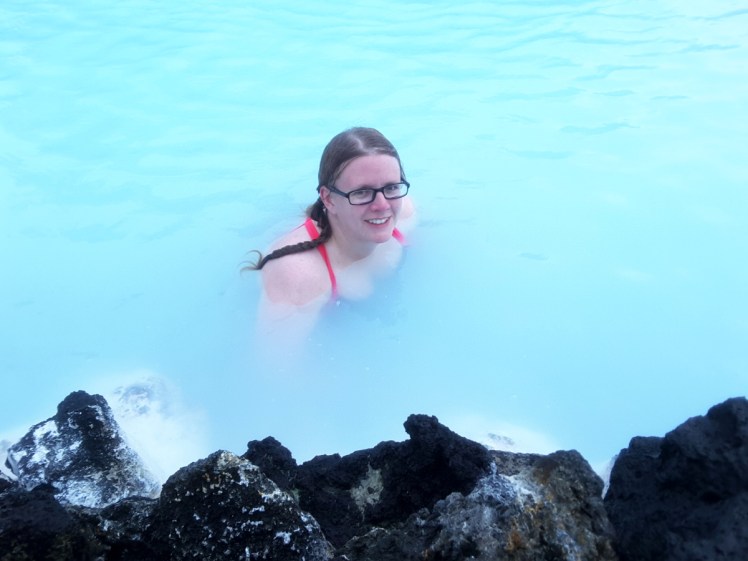 Greeter photo of me in the Blue Lagoon. The water is a really bright light blue and there are black lava rocks along the bottom of the picture.