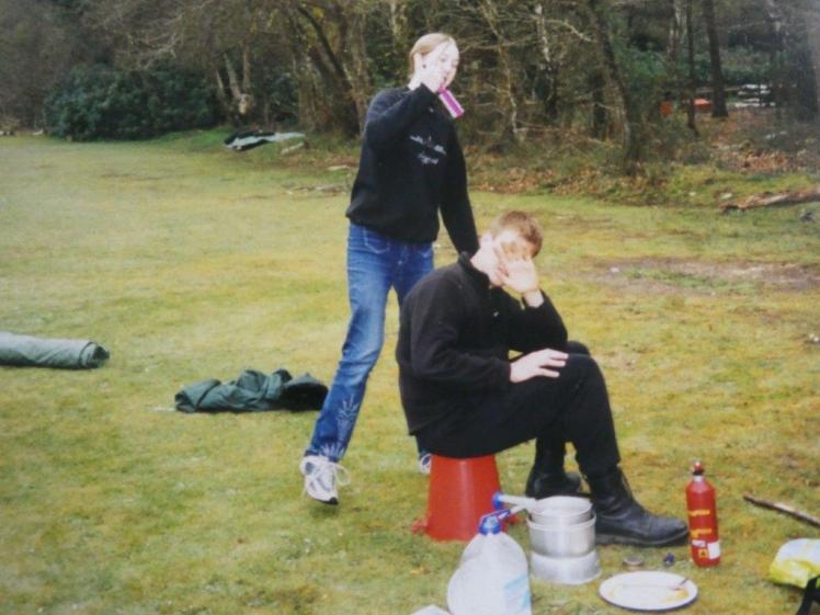Chris is sitting on the fire bucket by the stove with a hand over his face. Catherine is standing behind him pretending to stab him in the head with her hairbrush.