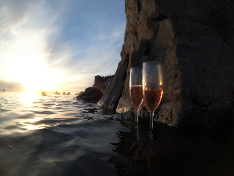 Two glasses of pink alcoholic fizz I found abandoned on a rock, with the low sun shining through them and reflecting on the water.