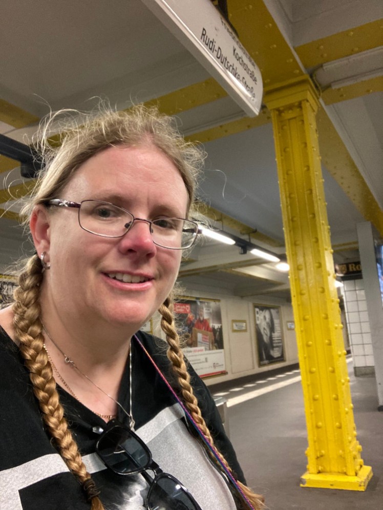 A selfie in the U-Bahn station. There's a yellow steel pillar holding the roof up and yellow supports running along the roof. The ads and station name are framed in yellow. I am not yellow, I am definitely pink and sweaty-looking.