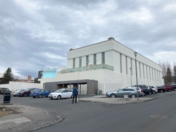 Sundhöllin, a pool in central Reykjavik. The original pool is housed in a white rectangular Art Deco building with frosted glass enclosing balconies halfway up the front. To the left is a new wall enclosing the outdoor extension and between the two is the glass wall of the lifeguard box, reflecting vivid blue pool water although at an angle that surely can't physically do that.
