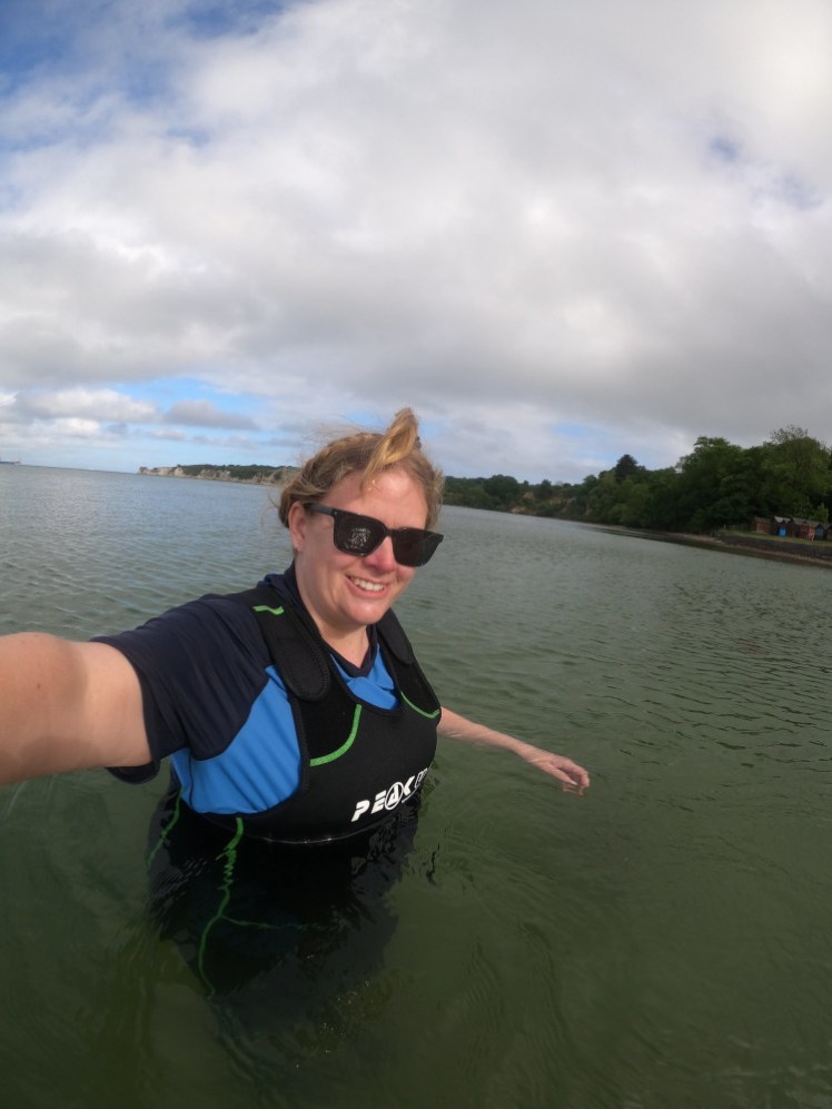 My first sea swim. I'm standing in grey-green water up to my waist. The logo on the front of the wetsuit is visible now. Rashvest as usual, plus sunglasses and I have my hair piled up on my head to keep it dry.