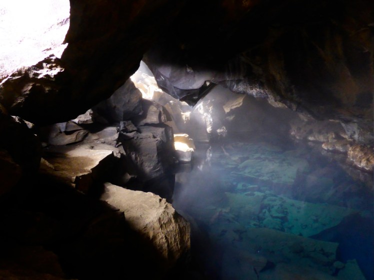 Inside a cave illuminated by shafts of sunlight coming through holes. The cave is absolutely littered with boulders, including under the surface of the bright blue steaming water.