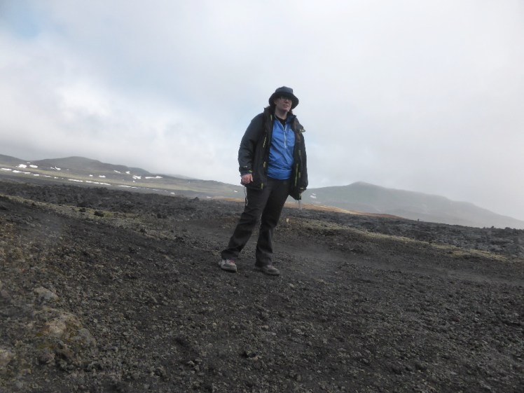 A timer selfie, wearing a blue windshirt under a black raincoat with bucket hat, on the black background of the Leirhnjukur lava field.