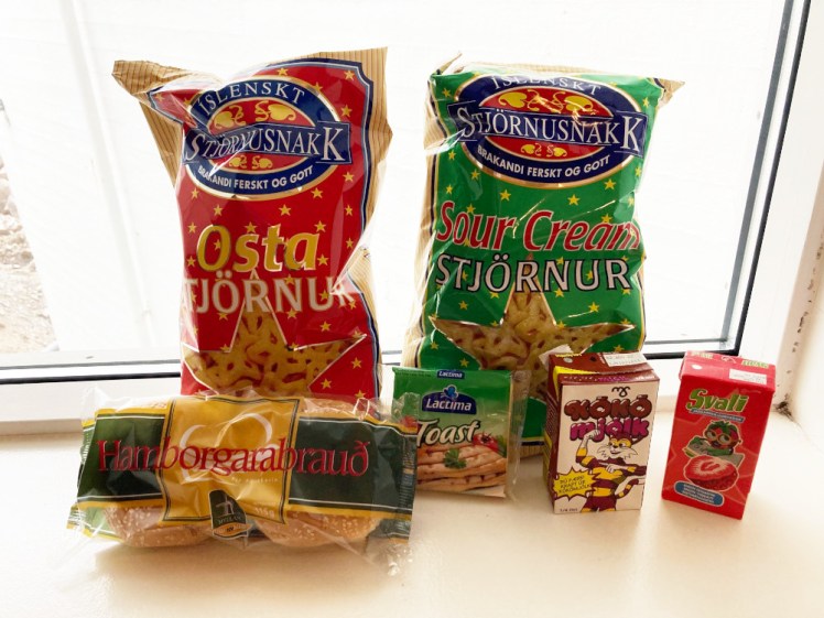 Two packs of star-shaped crisps. The red packet contains cheese flavour and the green packet contains sour cream flavour. There's also a packet of hamburger rolls, plastic cheese, a carton of chocolate milk and a carton of strawberry juice. These ones are sitting on the windowsill of my apartment in Reykjavik.
