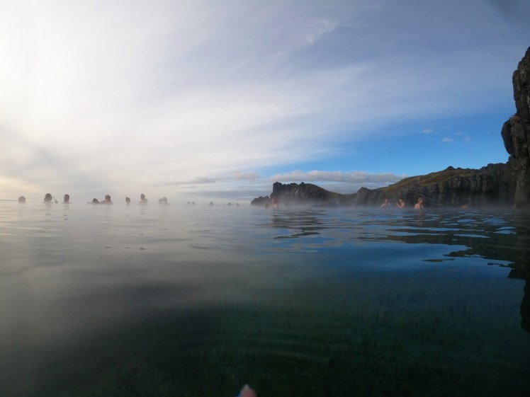 The sky lagoon from the south end. Part of the sky is blue, part streaked with clouds. At the end and to the right are low basalt cliffs and to the right, a line of people leaning on the infinity edge although from this distance you can't see the fjord beyond.