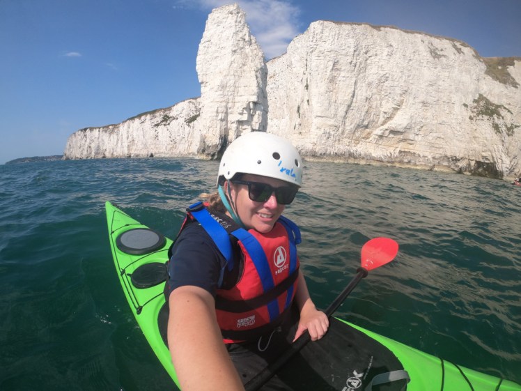 Me, in the long green kayak wearing a white helmet and a red buoyancy aid with blue straps. Behind me are the cliffs and a particularly spiky pinnacle.