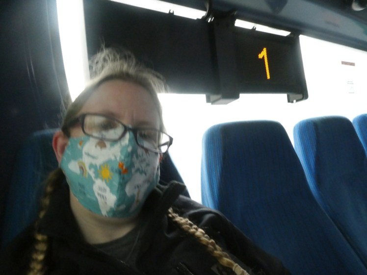 A selfie on a bus in Reykjavik. I'm wearing a light blue cloth mask with a map of the world on it. Behind me, the bus's LCD display says I'm on route 1, apparently.