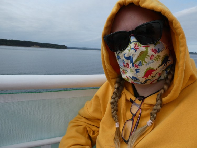 Selfie on the ferry, face hidden by mask, sunglasses and hood up. The mask has red, green & blue dinosaurs on it.
