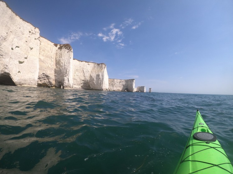 The nose of my long pointed green kayak on the water on the right. On the left is a line of white cliffs punctuated by towers and pinnacles. Old Harry is visible in the distance, all arches and stacks.