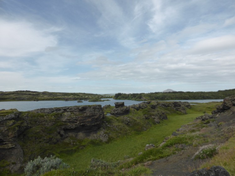 The lake shore at Skutastadir. It's green but it's also rocky and there are rocks sticking up as tiny islands in the water. On the other side you can see weird tiny humpy hills.