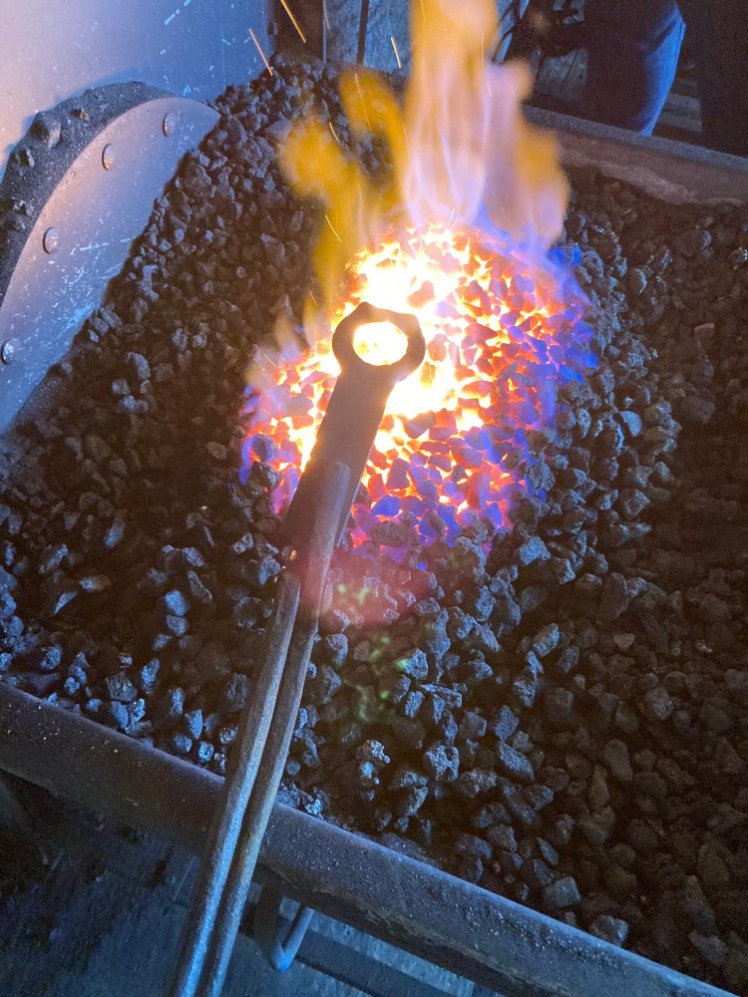 An iron bottle opener held in a long pair of tongs and held in the fire in the forge.