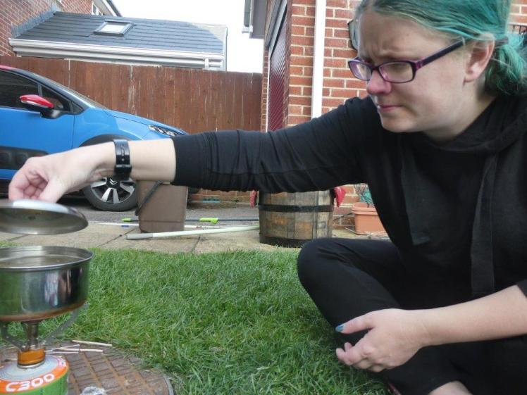 Cooking pasta on my camping stove. I've changed out of the white lacy dress and into black hoodie and leggings. My green hair is visible. I'm opening the pasta pot lid to check if the water's boiling yet.