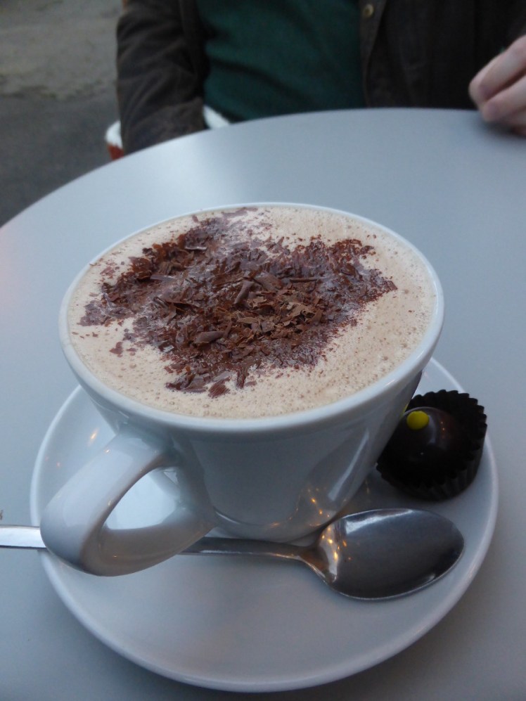Chococo orange hot chocolate, topped with foam and grated chocolate with a little chocolate in a miniature