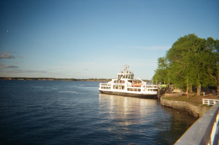 A ferry leaving Suomenlinna, seen from a bridge. Now the trees are on the right and you can see the ferry openly from the side.