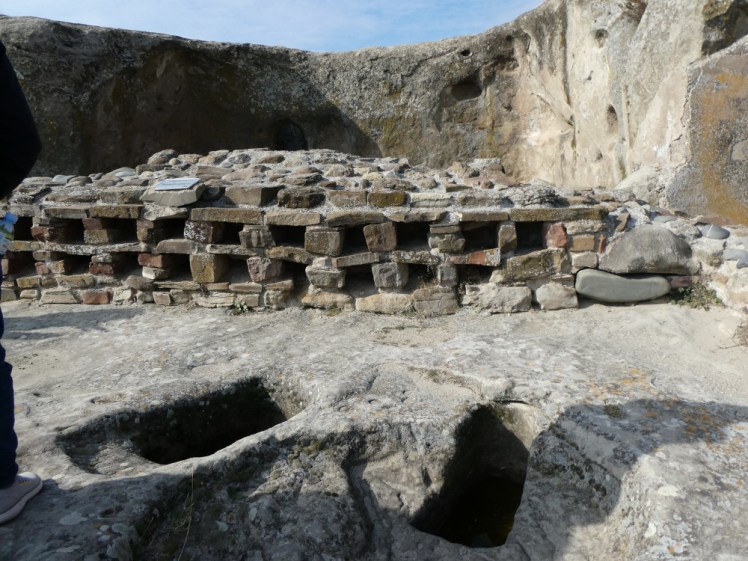 A reconstructed pharmacy with sets of open storage boxes rebuilt in the Soviet era. There are cold storage holes in the ground and a curved wall at the back.