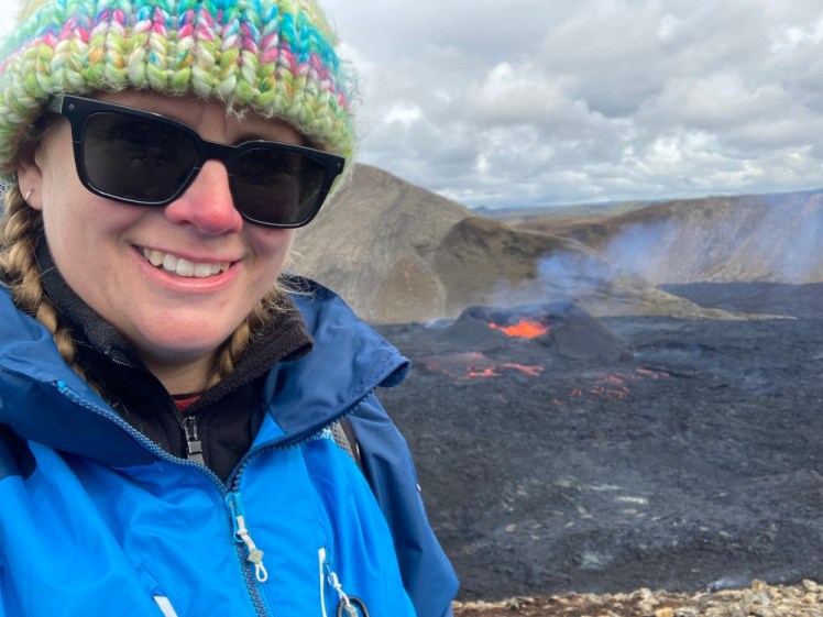 Selfie with a small active volcano, on a cloudy day. I'm wearing a bobbly hat and waterproofs and the volcano is happily splashing orange lava around.