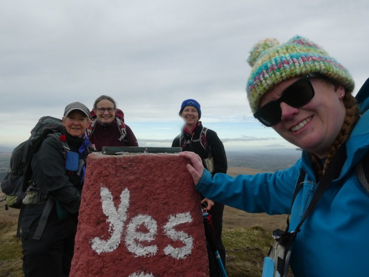 A group selfie at the trig point, which is painted red and says "Yes Cymru", apparently in support of the World Cup. The sky is grey now and we're all wearing a lot more clothes than in the earlier group photo.
