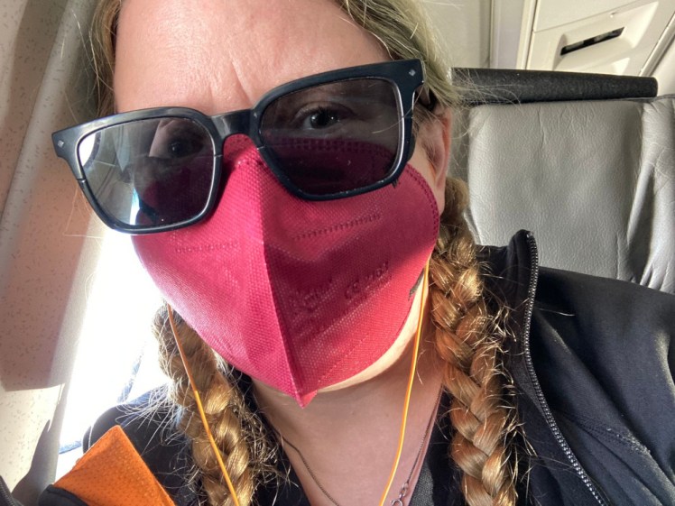 Me on a plane, wearing a red medical mask and sunglasses. You can clearly see my left eye but between the shadows and reflections, my right eye looks a bit like I've been punched.