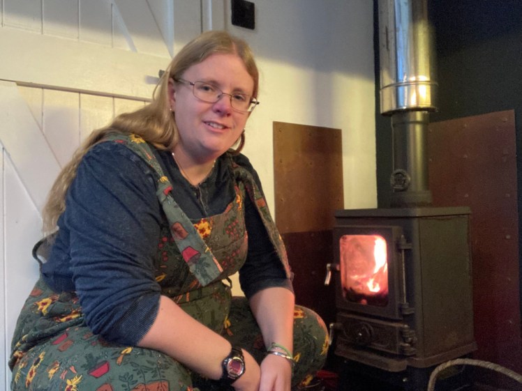 Crouched by the fire in green dungarees covered in gardening stuff and a long-sleeved denim-effect t-shirt.