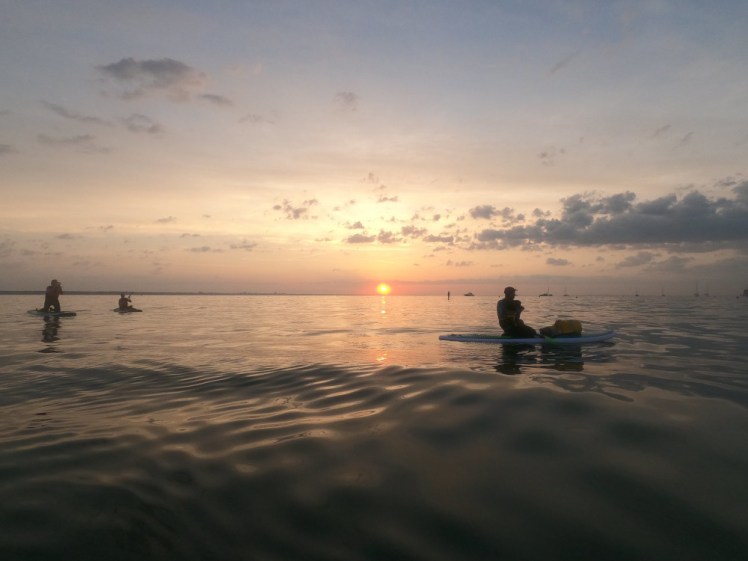 The sun centered just above the horizon, with the sky turning peach and purple. The group on their paddleboards are silhouetted on the calm sea.