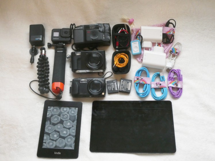 A GoPro on a floaty handle, an 80s film camera and two small Lumix digital cameras laid out beside various spare batteries, a tiny tripod, a tiny case open to show headphones and iPhone adaptor, two USB plugs and various cables tied up with Minnie Mouse wraps. At the bottom is my Kindle and my tablet, which, yes, does need a clean.