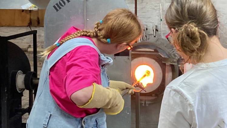 Me holding a lump of glass in the furnace on the end of a steel pole, watching intently as I turn it to make swirls.