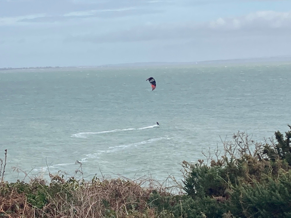 On a pale greenish sea against a sky only slightly more blue, a kitesurfer flies across the waves on a windy day.