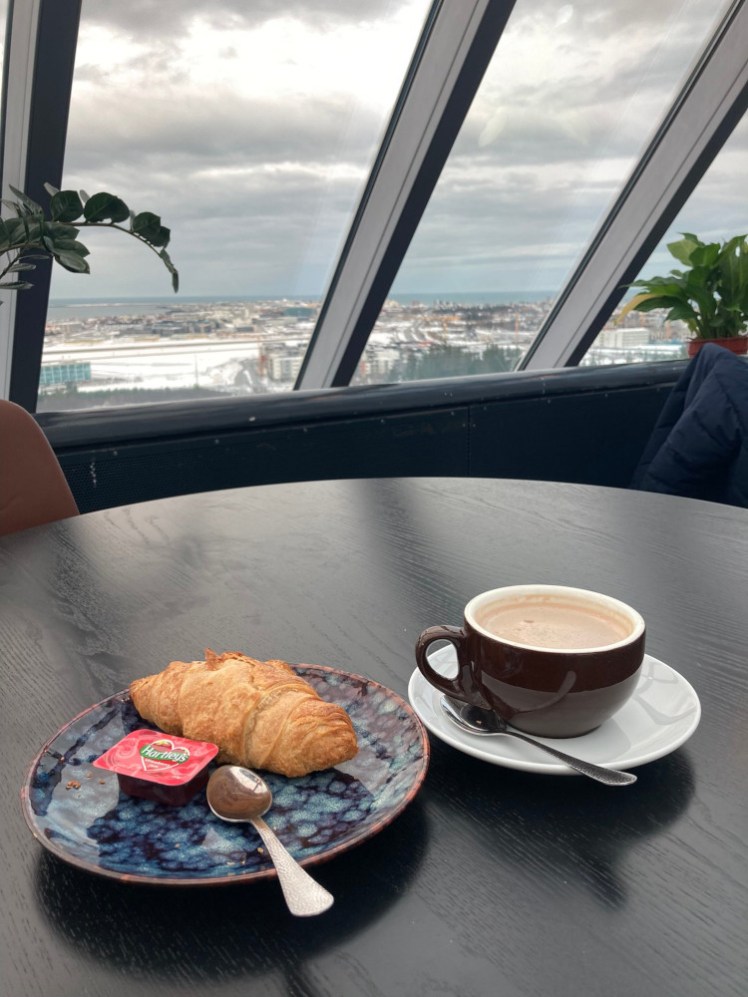 A croissant on a blue patterned plate and a cup of hot chocolate. Both are on a round dark table and behind the table is the curved edge of the huge glass dome roof.