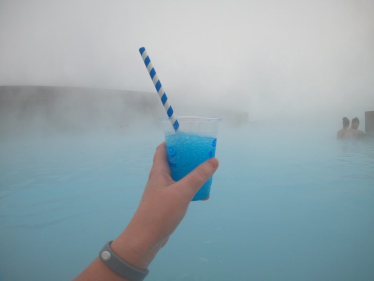 My arm sticking out of the Blue Lagoon, grey silicon wristband visible, holding up a reusable plastic glass of blue raspberry slushie with a paper straw sticking out of it.