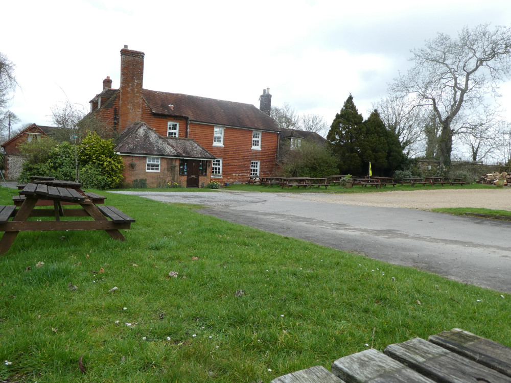 The Millburys, a slightly run-down-looking red brick pub as seen from a picnic bench halfway along the grass that borders the gravel car park.
